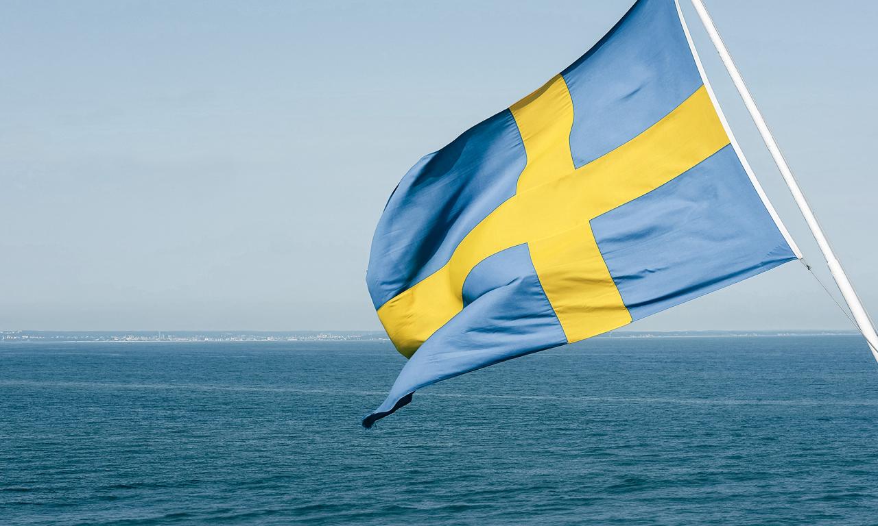 Svensk flagga som vajar i vinden med havet i bakgrunden.